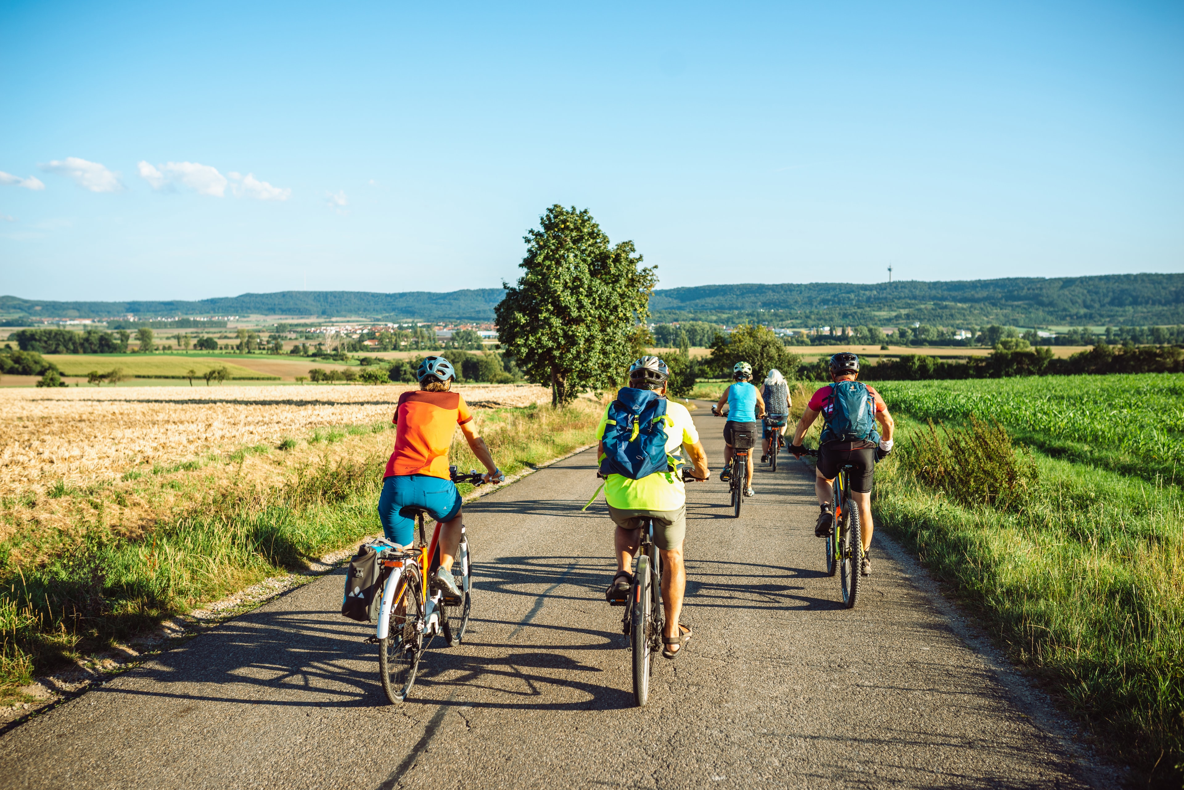 Mehrere Menschen mit Fahrrädern fahren auf einem hellen Schotterweg durch offene Landschaft mit Feldern und ein paar Bäumen.